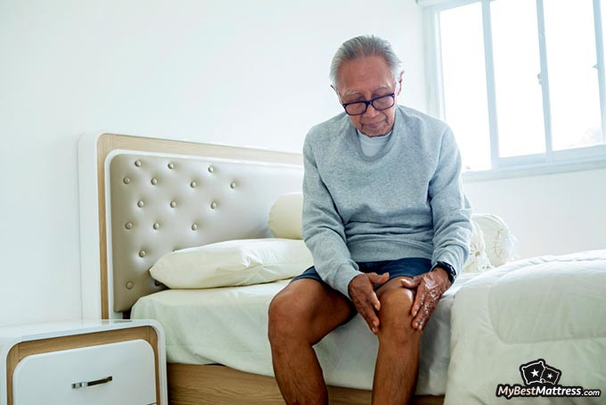 Knee pillow: a senior man holding his knee while sitting on a bed.